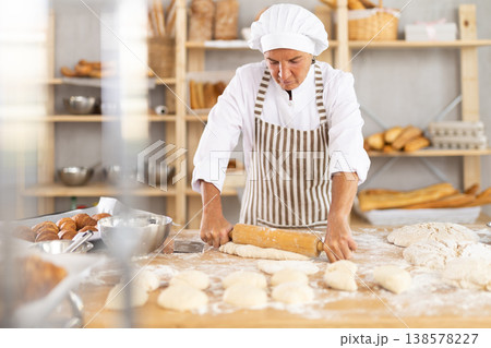 Aged woman baker using a rolling pin to flatten dough in a bakery Aged woman baker using a rolling pin to flatten dough in a bakery 138578227