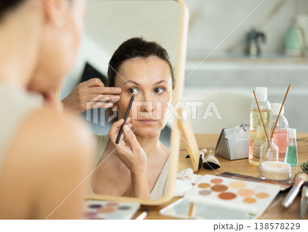 Woman reflected in tabletop mirror applying eyeliner during daily makeup 138578229