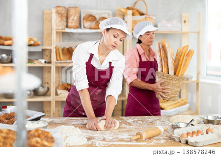 Two professional female baker in apron working in bakehouse. Young woman kneading dough and mature woman arranging freshly baked bakery products Two professional female baker in apron working in bakehouse. Young woman kneading dough and mature woman arranging freshly baked bakery products 138578246