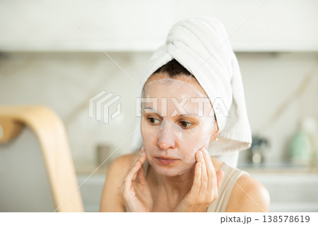 Middle-aged woman applying tissue mask on her facial skin sitting in front of the mirror Middle-aged woman applying tissue mask on her facial skin sitting in front of the mirror 138578619