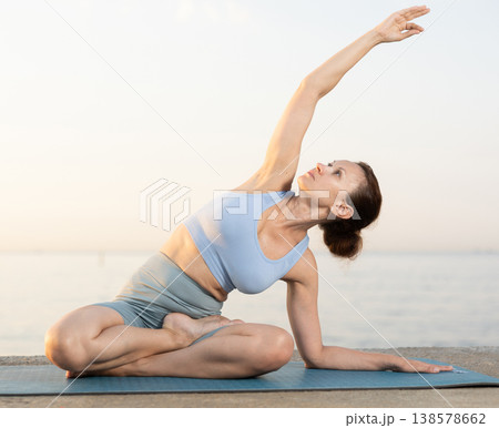 Sporty energetic female doing exercises parshvottanasana pose of yoga sitting on pilates mat during training on beach at sunrise Sporty energetic female doing exercises parshvottanasana pose of yoga sitting on pilates mat during training on beach at sunrise 138578662