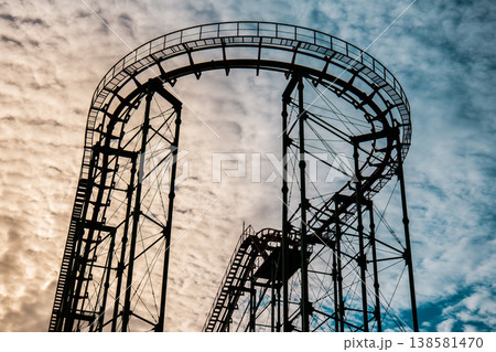 Dark silhouette of a roller coaster track against a bright, textured cloud background at dusk Dark silhouette of a roller coaster track against a bright, textured cloud background at dusk 138581470
