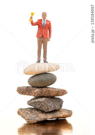 Miniature person stands on stacked stones while holding a trophy in a bright space during the day 138584097