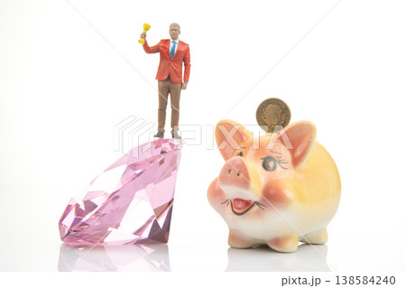 Miniature person holds trophy while standing on gem next to piggy bank with coin Miniature person holds trophy while standing on gem next to piggy bank with coin 138584240
