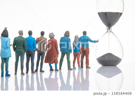 Miniature figurines of people gathered around an hourglass at a neutral background during daytime Miniature figurines of people gathered around an hourglass at a neutral background during daytime 138584409