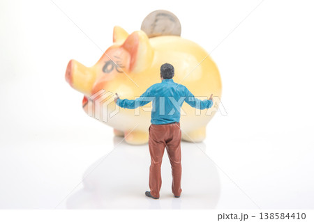 Miniature person stands near a large piggy bank with arms raised while facing the bank  138584410