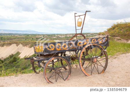 Old cart rests on hills overlooking a valley with green fields and distant mountains on a cloudy day 138584679