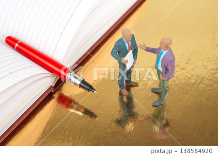 Miniature figurines have a discussion next to a large pen and an open planner on a shiny surface Miniature figurines have a discussion next to a large pen and an open planner on a shiny surface 138584920