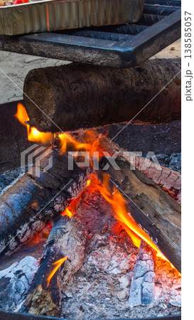 Campfire with burning logs and glowing embers, metal grill rack above, outdoor setting with ash and smoke visible in the background 138585057
