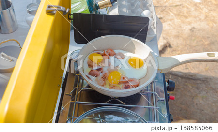 Cooking breakfast outdoors with eggs and sausages in a frying pan on a portable camping stove in a natural setting 138585066