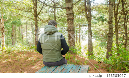 Man sitting on a wooden bench in a forest, wearing a green vest and cap, surrounded by trees and a view of a lake in the distance 138585067