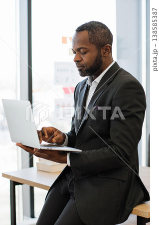 An African American man in a suit sits at a desk, focused on his laptop, with a large window behind him 138585387