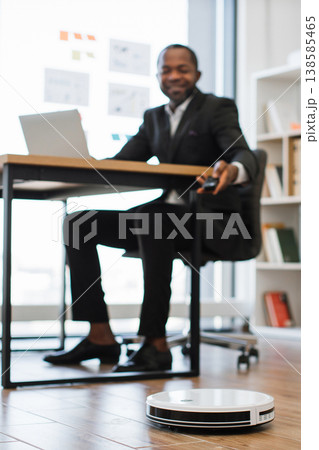 An African American man in a suit smiles while working on a laptop at his desk as a robot vacuum cleaner operates on the floor An African American man in a suit smiles while working on a laptop at his desk as a robot vacuum cleaner operates on the floor 138585465