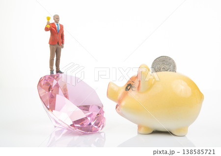 Miniature people stand on a crystal holding a trophy, while a piggy bank waits with a coin resting  138585721