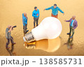 Miniature people gather around large light bulb on shiny golden surface during a creative discussion 138585731