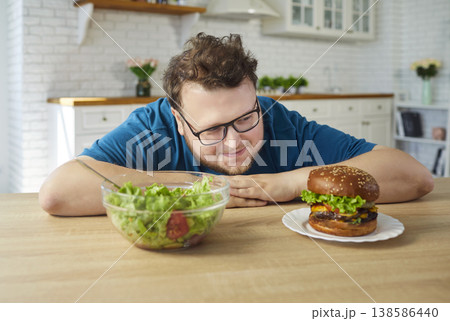 Obese man choosing salad or juicy burger at home kitchen, diet temptation Obese man choosing salad or juicy burger at home kitchen, diet temptation 138586440