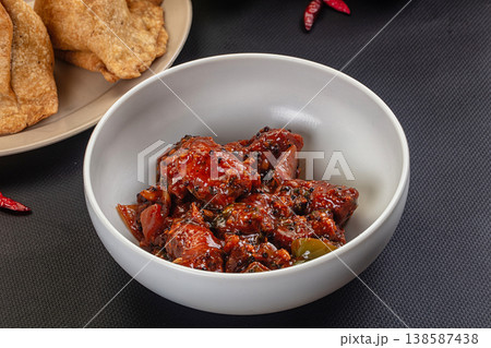 Spicy Chicken Indo-Chinese appetizer with a rich, glossy red sauce on a beige plate, set on a dark tabletop. 138587438