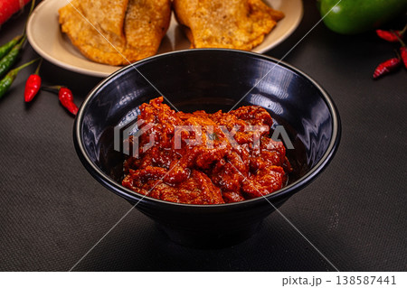 Spicy red chili pickle or chutney in a white bowl with fried snacks and fresh vegetables. 138587441