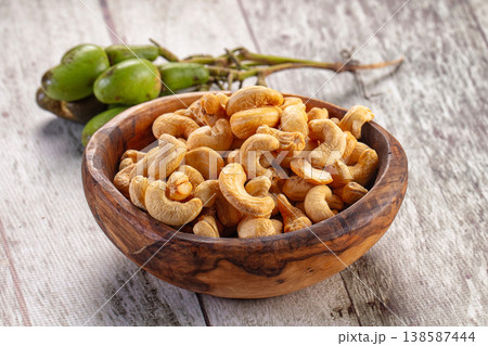 Roasted cashew nuts in a brown bowl with a cluster of raw green cashew fruit on a rustic wooden table. Roasted cashew nuts in a brown bowl with a cluster of raw green cashew fruit on a rustic wooden table. 138587444