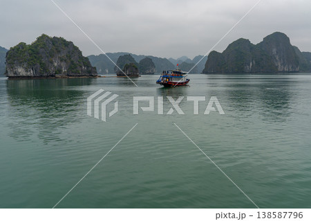A tourist boat navigates through the iconic limestone karsts of Ha Long Bay under an overcast sky A tourist boat navigates through the iconic limestone karsts of Ha Long Bay under an overcast sky 138587796