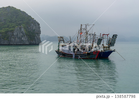Two traditional Vietnamese fishing vessels are anchored in the calm waters of Ha Long Bay amidst a misty morning 138587798