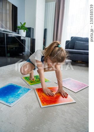 Little girl kneeling on floor pressing colorful sensory gel mats with hands during play activity. Sensory stimulation, autism support and occupational therapy development concept. Little girl kneeling on floor pressing colorful sensory gel mats with hands during play activity. Sensory stimulation, autism support and occupational therapy development concept. 138588539