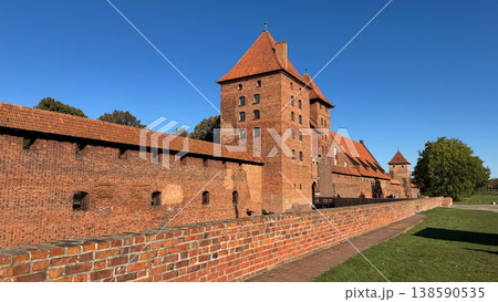 Malbork Castle in Poland with red brick walls towers and long defensive fortifications under clear blue sky viewed along pathway and grass. Architecture, history and travel concept Malbork Castle in Poland with red brick walls towers and long defensive fortifications under clear blue sky viewed along pathway and grass. Architecture, history and travel concept 138590535