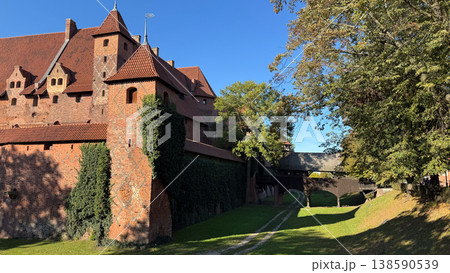 Malbork Castle in Poland with red brick walls tower and wooden bridge over grassy moat with trees under blue sky. Architecture, history and travel concept 138590539