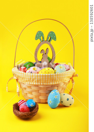Colorful hand-painted Easter eggs in a wicker basket with a wooden bunny ornament and a small nest against a vibrant yellow background. Cheerful spring holiday decoration. High quality photo 138592601