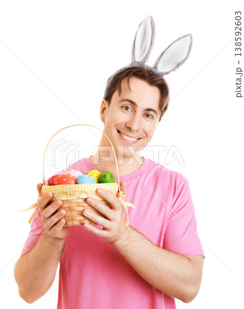 Smiling young man wearing bunny ears and a pink t-shirt holding a basket full of colorful painted Easter eggs. Isolated on a white background. Cheerful spring holiday portrait. High quality photo 138592603