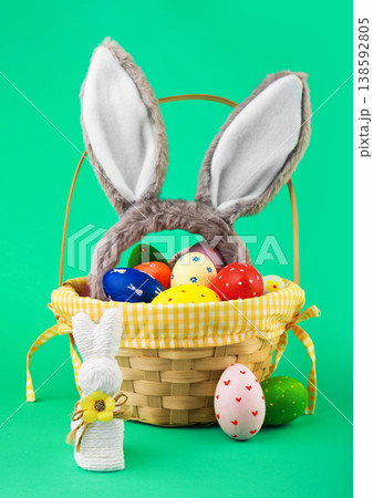 Festive Easter basket filled with colorful painted eggs and fluffy bunny ears. A white string bunny and decorated eggs sit nearby against a bright green background. High quality photo 138592805