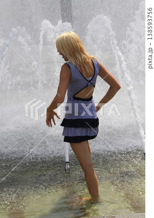 Young blonde hair woman, stands barefoot in a shallow fountain,,surrounded by splashing water jets. Young blonde hair woman, stands barefoot in a shallow fountain,,surrounded by splashing water jets. 138593756