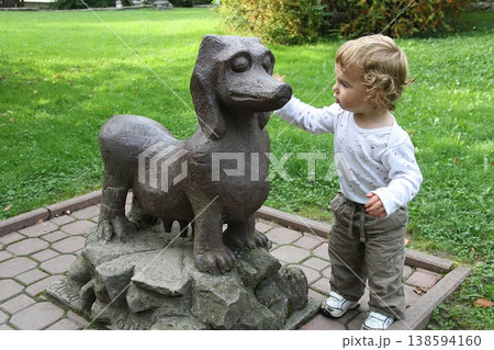 Curious toddler gently touches a small dog statue in a lush park. Curious toddler gently touches a small dog statue in a lush park. 138594160