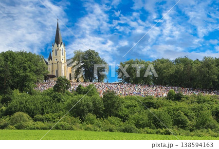 Large crowd gathers on a hillside near a tall church under a vibrant blue sky. Large crowd gathers on a hillside near a tall church under a vibrant blue sky. 138594163
