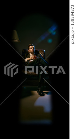 Elegant man drinking whiskey in armchair under dramatic lighting seen through keyhole. 138594873