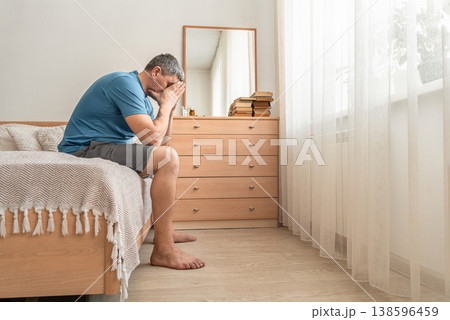 Man sitting on bed, covering face with hands showing signs of distress, loss, and mental health issues. Mental health of middle-aged people, midlife crisis 138596459