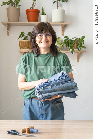 Smiling adult woman standing, holding a stack of old denim jeans for cutting and sewing, promoting sustainable clothing reuse. Environmental awareness Smiling adult woman standing, holding a stack of old denim jeans for cutting and sewing, promoting sustainable clothing reuse. Environmental awareness 138596513