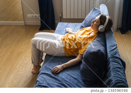 Woman lying on couch wearing white wireless headphones, enjoying audio from smartphone, surfing the internet in a cozy living room Woman lying on couch wearing white wireless headphones, enjoying audio from smartphone, surfing the internet in a cozy living room 138596598
