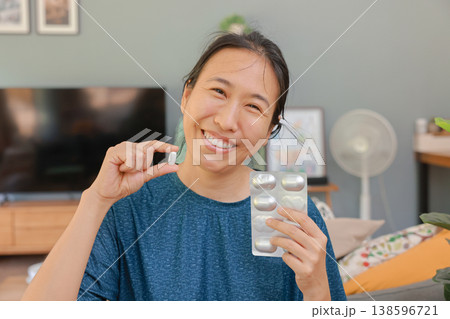 Happy Asian woman holding a white pill and blister pack at home. Healthcare, vitamin, supplement, or pain relief concept. Smiling patient recovered from illness showing medication to camera. 138596721