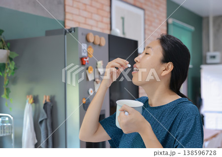 Side profile of young Asian woman putting a white pill into her open mouth and holding a blister pack. Taking daily medicine, vitamin, painkiller or dietary supplement for medical healthcare concept. Side profile of young Asian woman putting a white pill into her open mouth and holding a blister pack. Taking daily medicine, vitamin, painkiller or dietary supplement for medical healthcare concept. 138596728