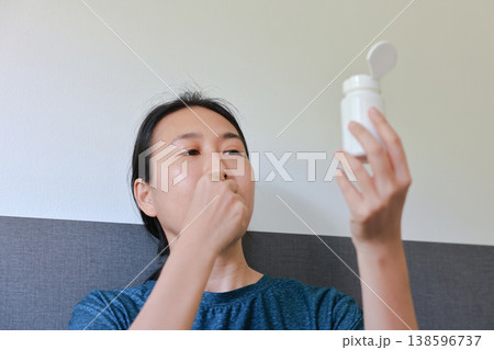 Pensive Asian woman holding and inspecting an unlabeled white pill bottle. Healthcare routine, medication management, dosage check, and concern about treatment concept. Pensive Asian woman holding and inspecting an unlabeled white pill bottle. Healthcare routine, medication management, dosage check, and concern about treatment concept. 138596737