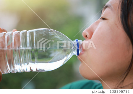 Close up side view of young Asian woman drinking pure mineral water from a clear plastic bottle outdoors. Healthy hydration, quenching thirst, wellness, body refreshment and summer lifestyle concept. 138596752