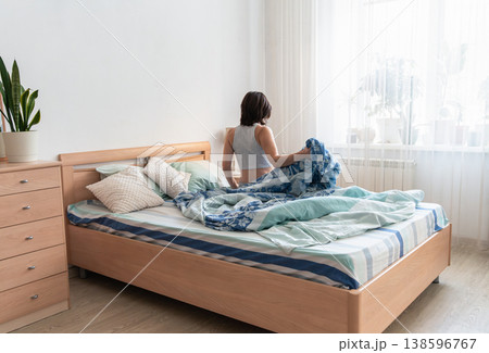 Woman sitting up in bed in a bright bedroom, starting the morning and looking towards the daylight from the window. The ordinary life of a real person. Authentic lifestyle 138596767