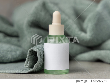 Glass dropper bottle with blank label mockup near sage green bath towel, unbranded packaging 138597769