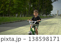 Three year old boy smiles and looks at the camera while riding a green kids bicycle on asphalt in the park with a forest background 138598171