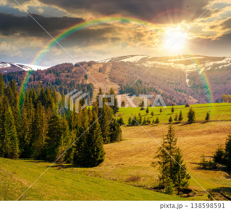 sunset over mountain landscape during spring. peaceful countryside scenery with coniferous forest near grassy field. beautiful ridge with snow capped top in the distance under the rainbow 138598591