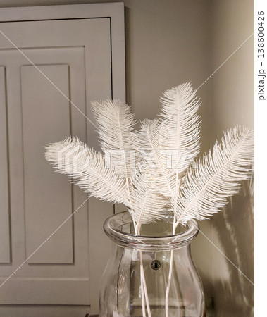 White feather decoration in a glass vase set against a neutral wall background with soft lighting highlighting the texture and elegance of home decor elements 138600426