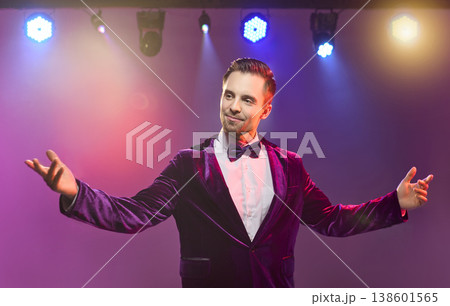 Charismatic show host on stage under colorful spotlights, welcoming audience 138601565