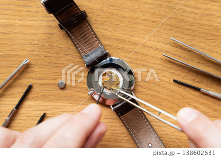 Close-up of a watch repair technician using tweezers to adjust the internal mechanism of a wristwatch on a wooden work surface Close-up of a watch repair technician using tweezers to adjust the internal mechanism of a wristwatch on a wooden work surface 138602631