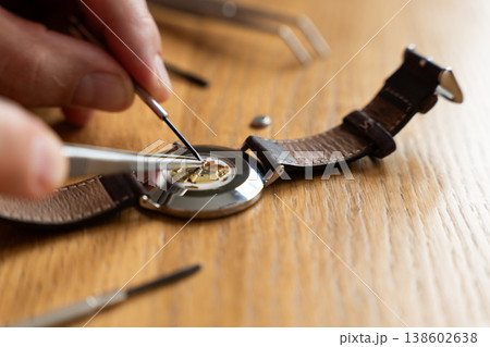 Close-up of a person using tweezers to repair a watch movement on a wooden workbench, with tools visible in the background Close-up of a person using tweezers to repair a watch movement on a wooden workbench, with tools visible in the background 138602638